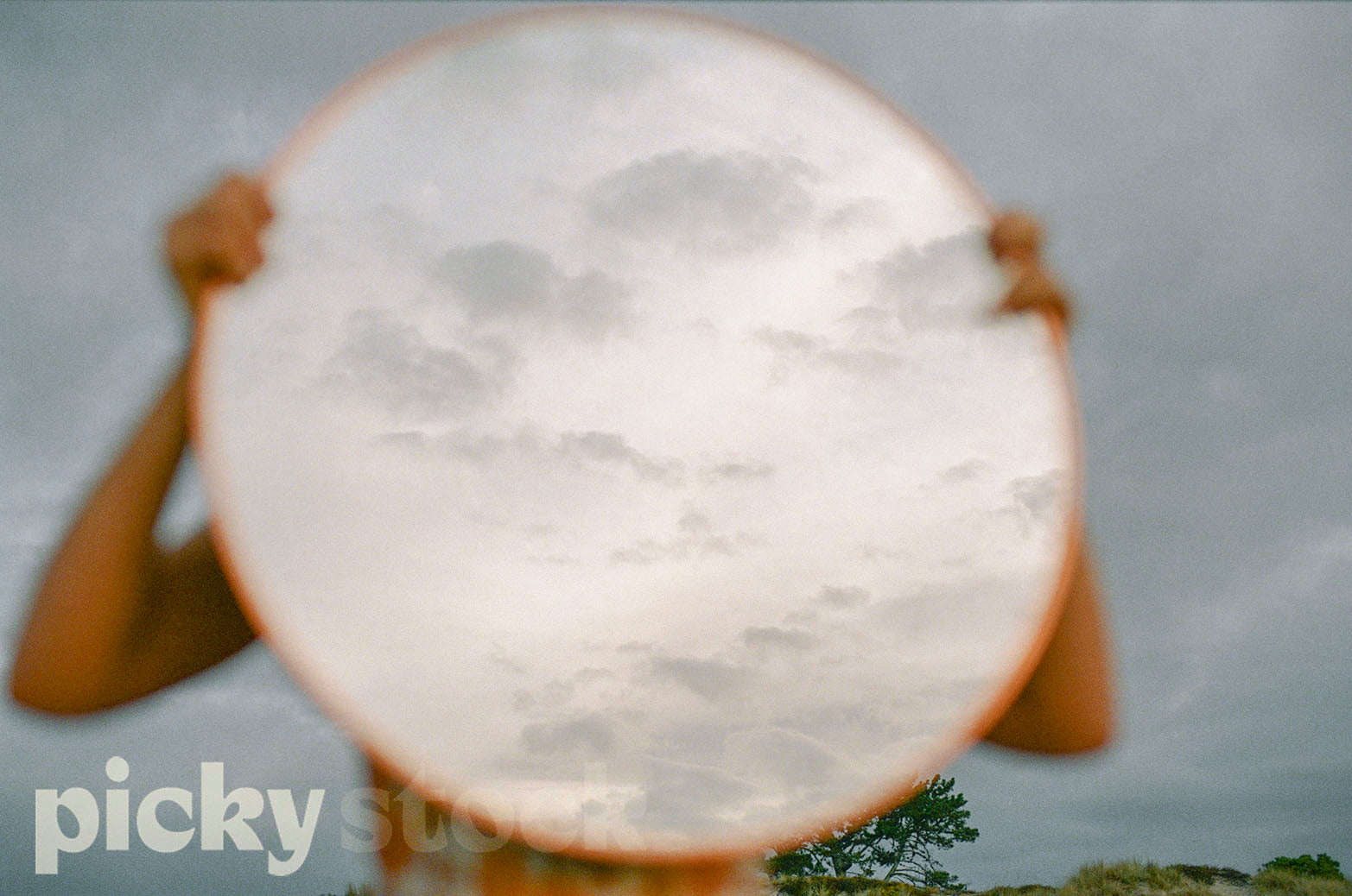 Lady holding a large round mirror towards the sky. Mirror is reflecting a soft white tone, with clouds. Background is a soft evening sky with green trees in the fair background. 