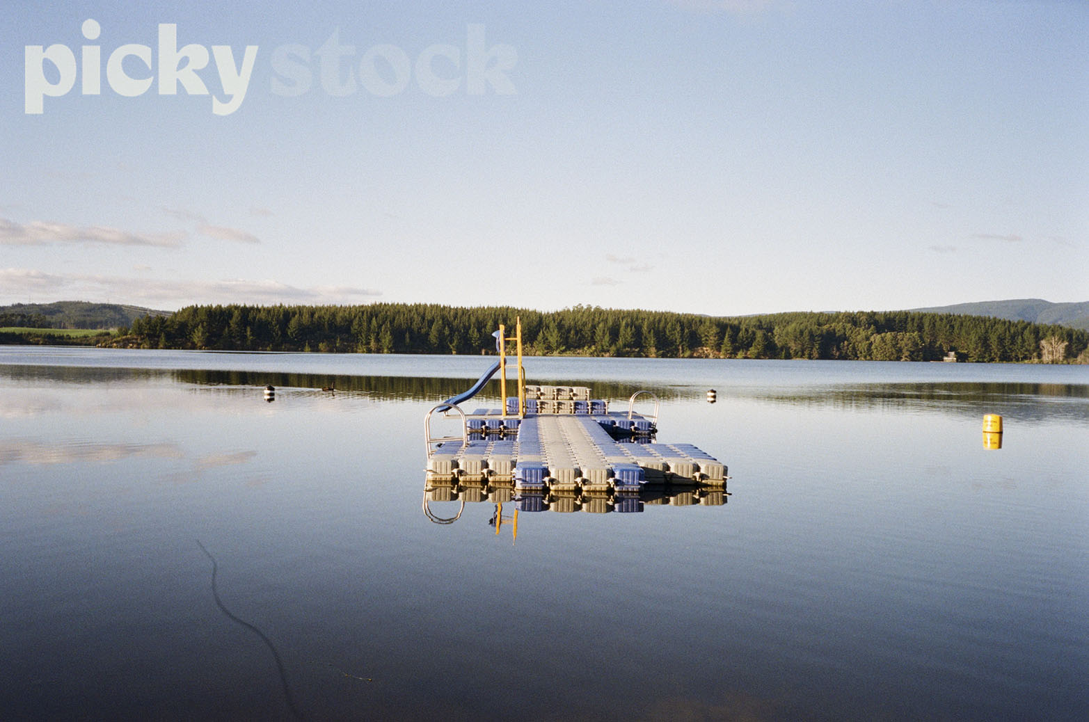 Floating pontoon in middle of river. Yellow ladder with blue slide attached. Ocean is calm, and a soft blue. Sky has a few scattered clouds, with green trees and bush at the other end of the lake. 