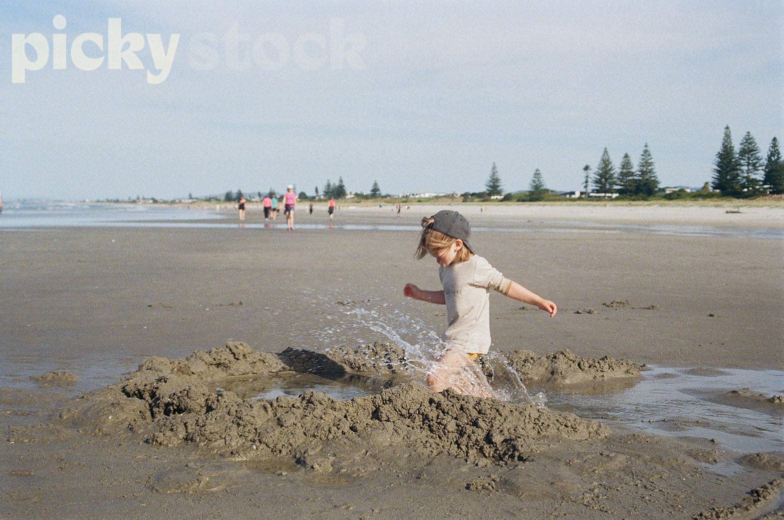 Image of young boy running through sandcastle with water splashing. Kid is wearing a cap, with mid length hair. Bunch of people running behind him. Ocean in background. 