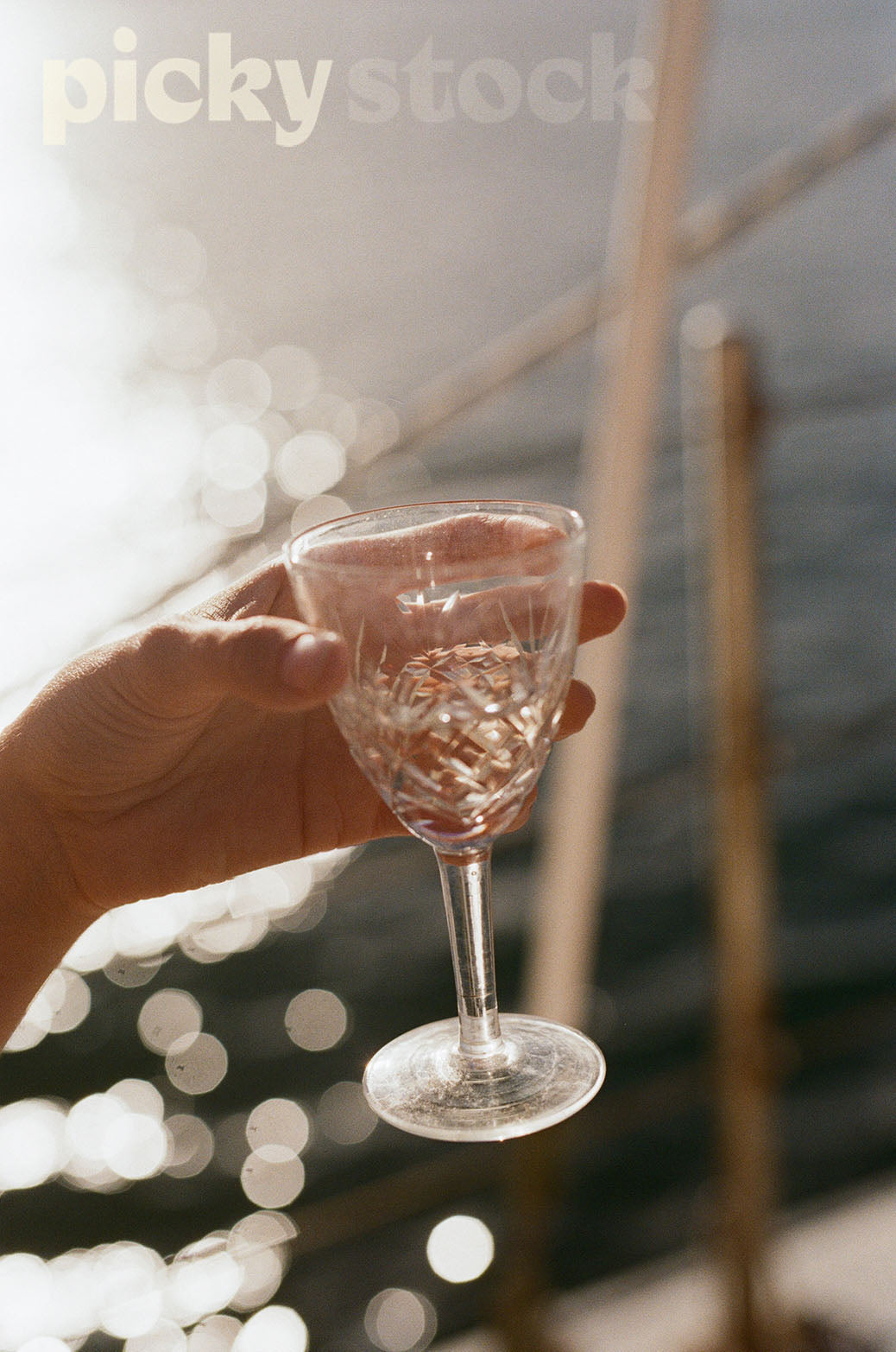 Lady holding a crystal champagne glass. Bokeh in the background against soft ocean water. Ladys hand in view only, background against a sailing boat.