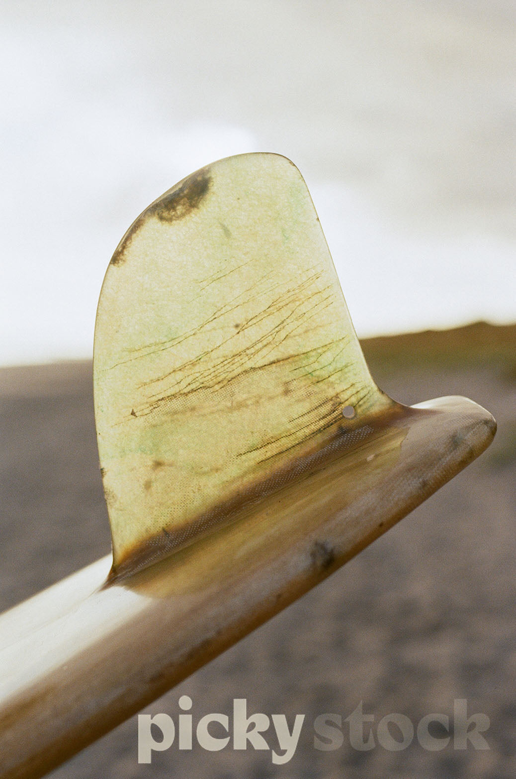 Close up image of surf board wide tail. Scratches and small marks are visible on the tail. Background is blown out sand and sjy