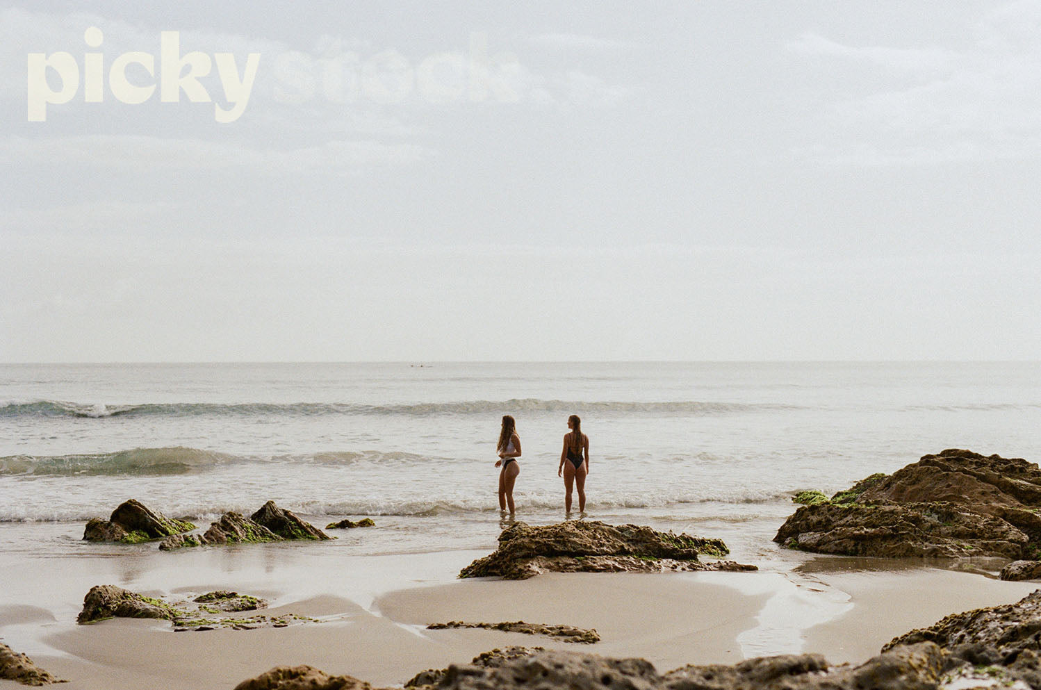Two girls standing in swim suits looking out to the ocean. Sea has a few waves, but relatively calm. Standing next to large rocks, at low tide. 