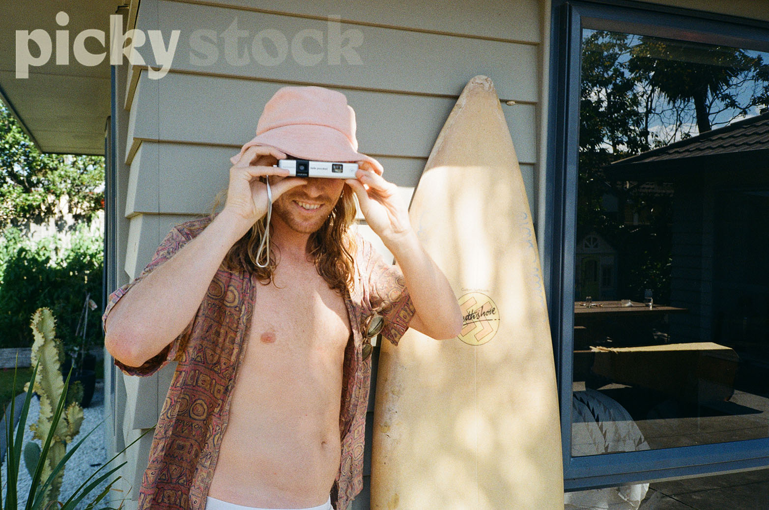 Man wearing a pink bucket hat holding a small film camera, taking a photo, eyes to camera. Man is wearing a patterned casual shirt with the buttons undone with bare chest underneath. Behind him is house, looks like a holiday home with a yellow surf board standing up to the right of him. 