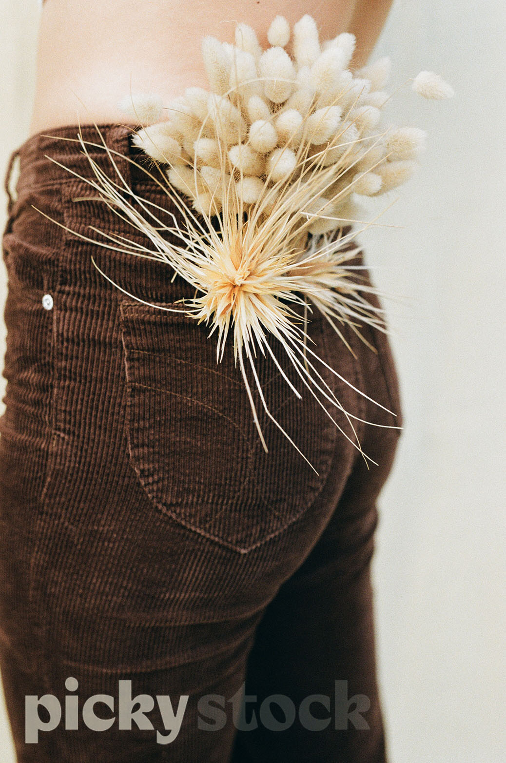 Close up of a pair of brown cord pants, with bunny tails and dried flowers in the back pocket. Image is cropped at the waist. Person is not wearing a top. Background is white / neutral. 