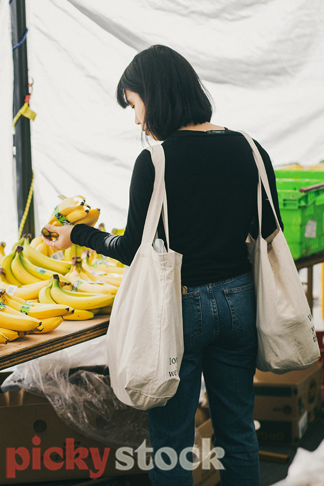 Young woman shopping for produce farmers market with reusable tote bags buying fair trade bananas.