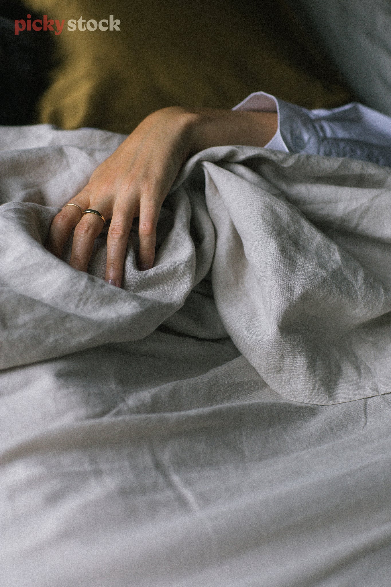 Unrecognisable woman lies in bed amongst unmade light grey sheets, her hand grips the sheets. 