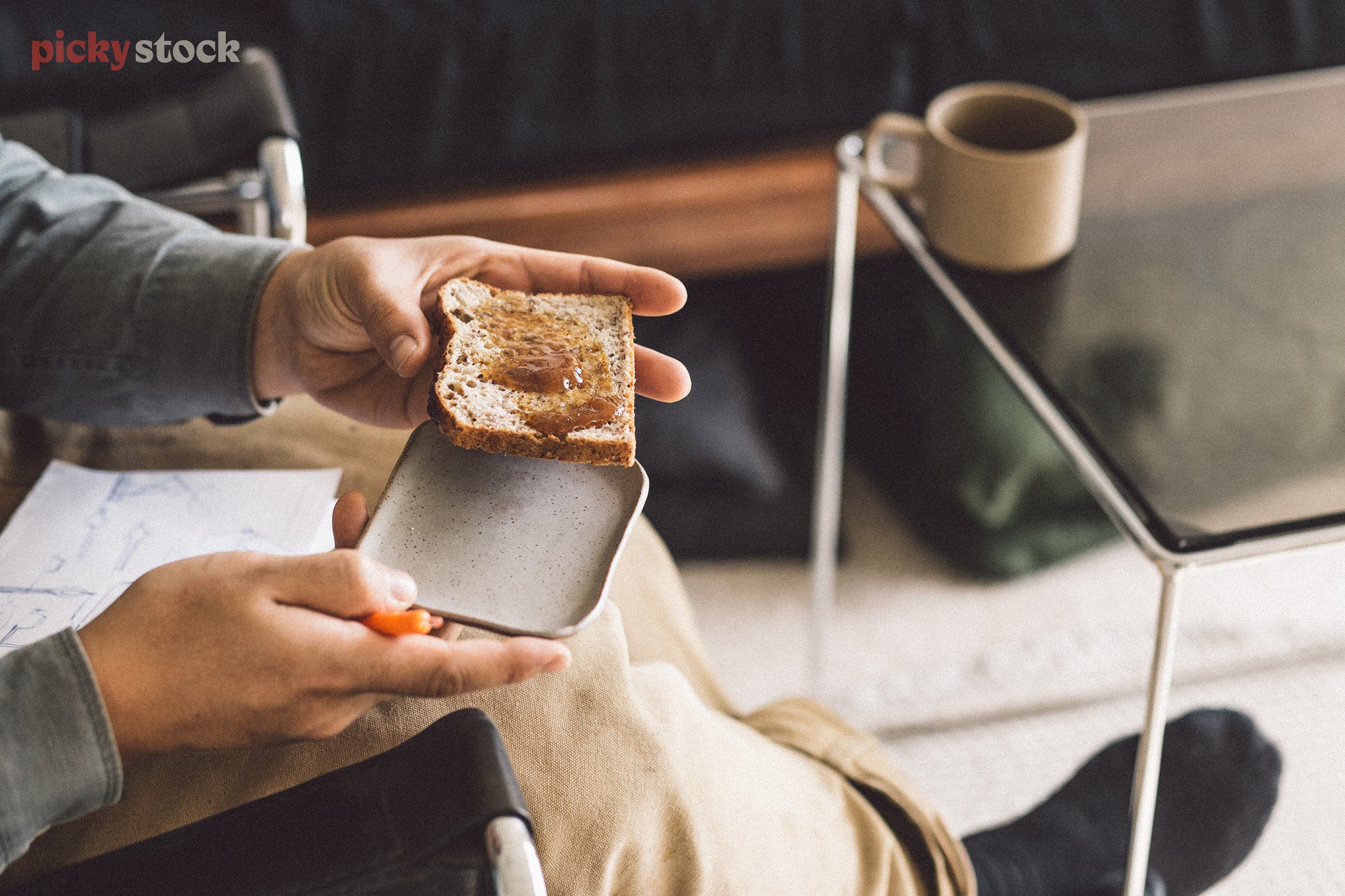 Unrecognisable man holds plate and jam on toast, while sitting near coffee table. 