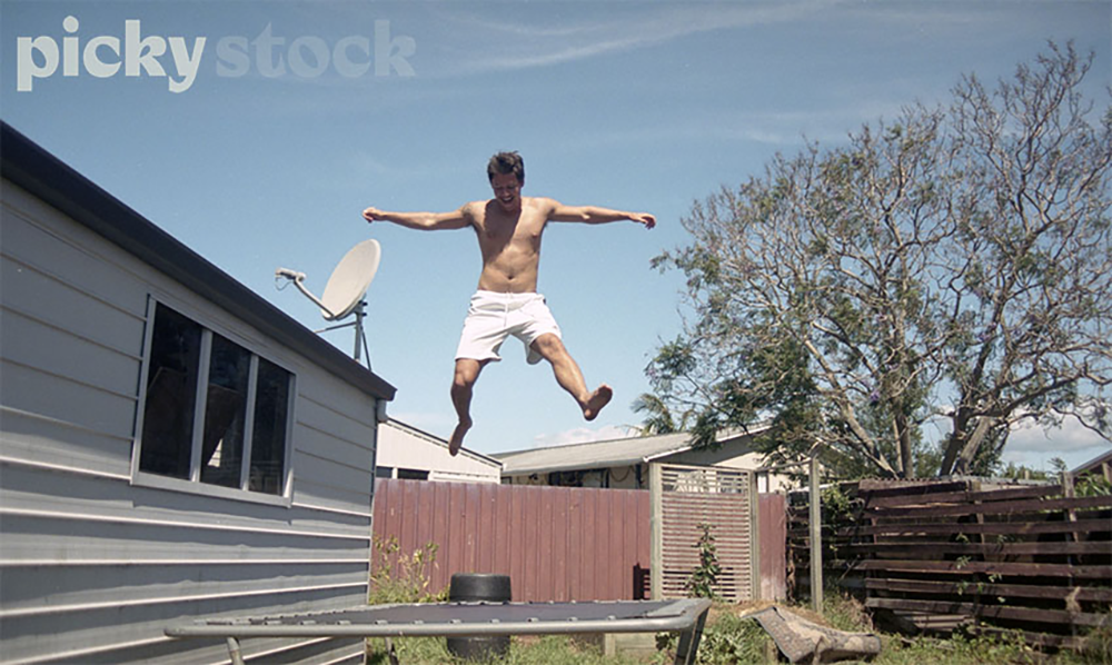 Young man jumps high on trampoline in back yard next to garage.