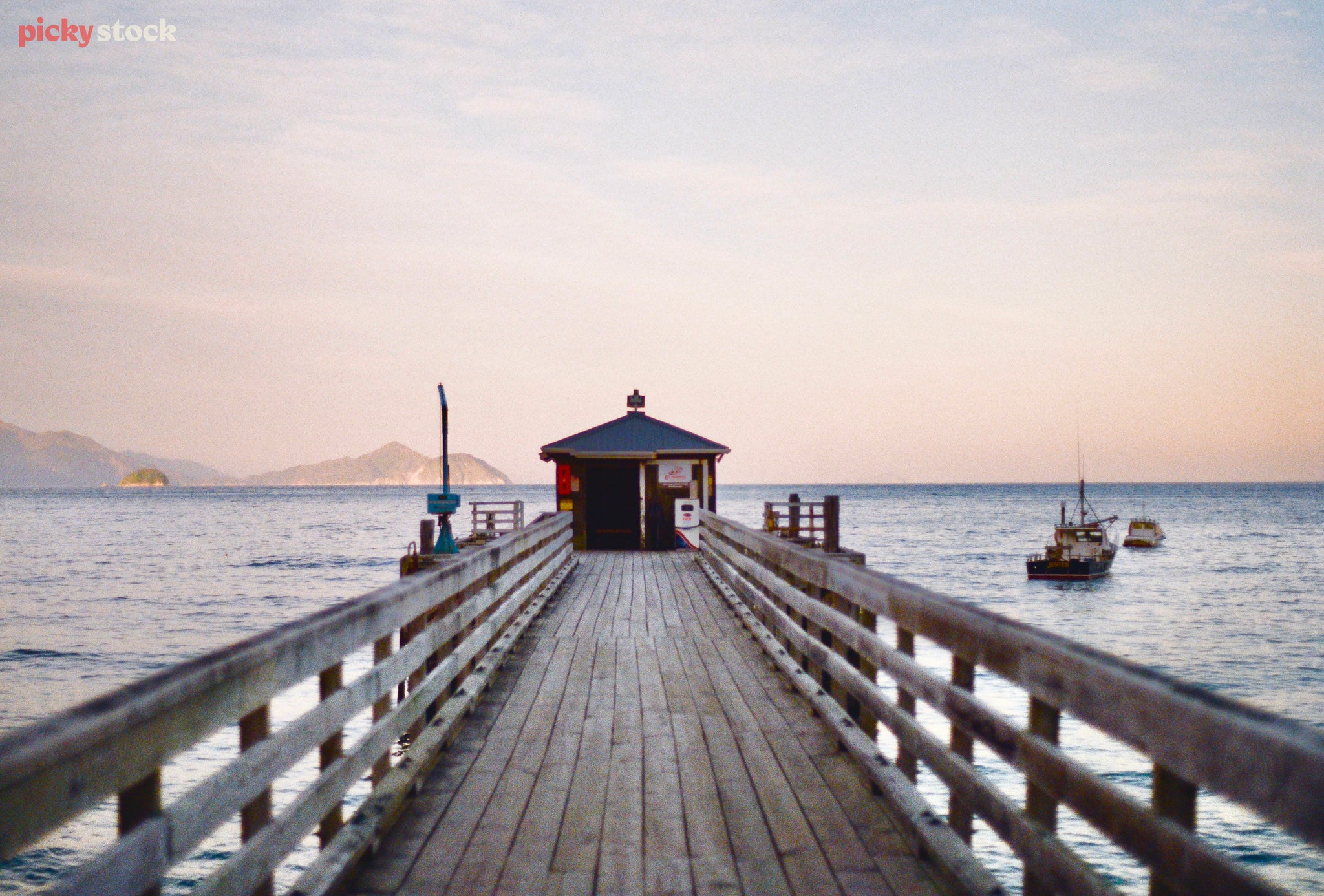 image of French Pass Pier at sunset. Boats to the right of the image in the sea. Water is blue, light is golden, soft sunrise. Small green building at the end of the pier 