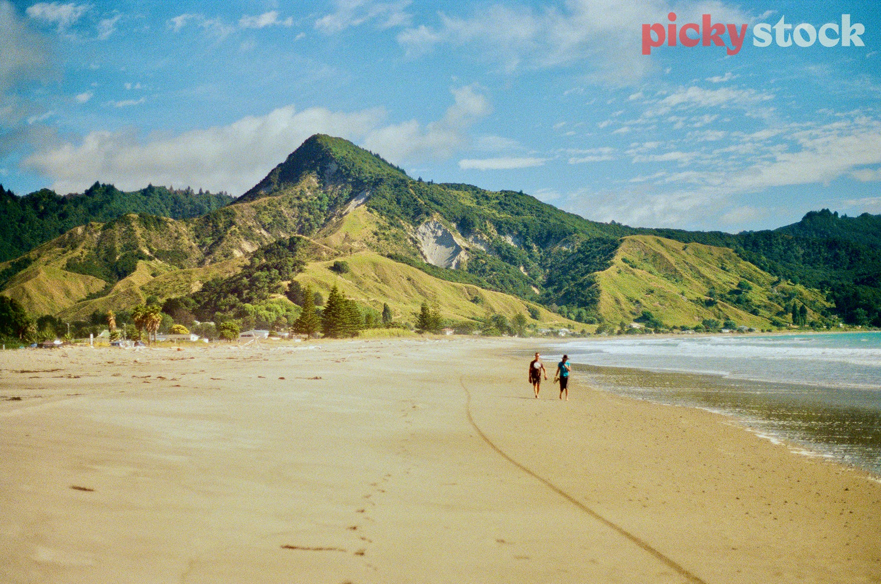 Wide landscape shot of two people walking down the beach, facing camera. Low tide with dramatic hills in background, and small settlement, campsite at the edge of the sand. Sky is blue with scattered clouds
