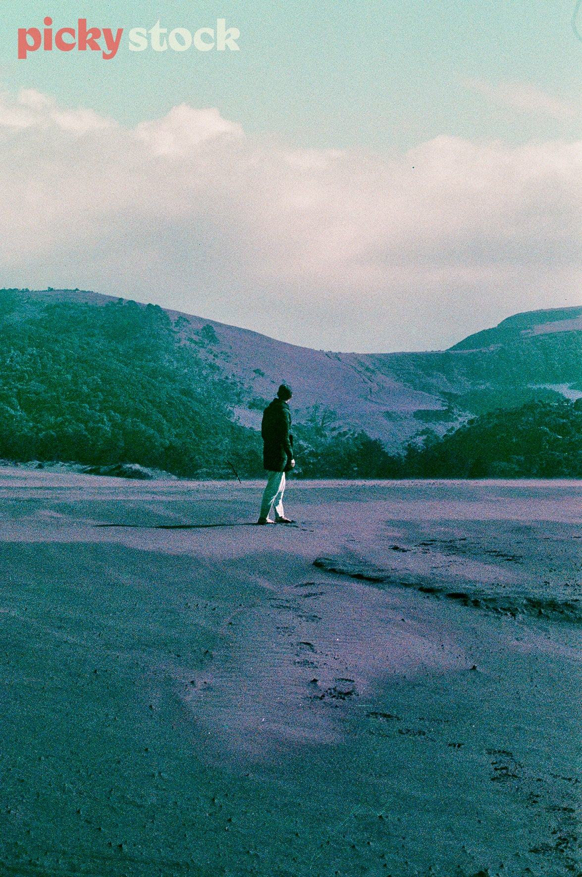 Film saturated image of man wearing beanie standing in sand looking out to mountains, Image has a blue grade across image