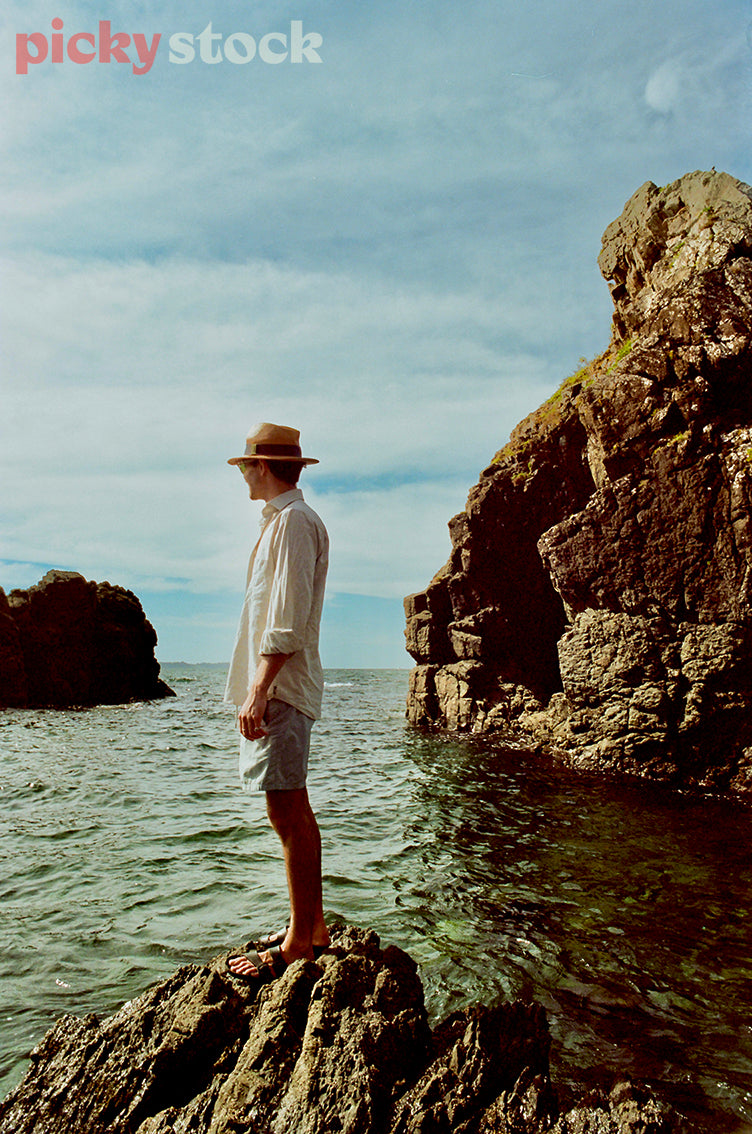 Man wearing a hat, white shirt and jean shorts standing on rocks wearing jandals. Overlooking the ocean. Other large rock formations in the background. Sky is a soft blue with a few full clouds. Water / sea / ocean is calm with a green tint. 