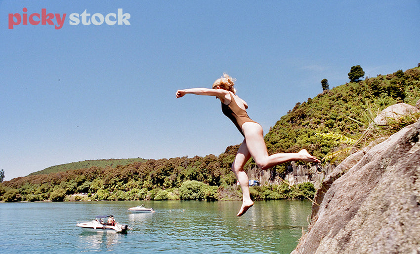 Woman mid-jump, mid flight from a large rock formation falling into Lake Taupō. Water is a blue and green colour. Scene is middle of the day. Lady is wearing a brown onepiece swim suit with barefeet. Green hills in the background and cliff facing. Two small boats in the background