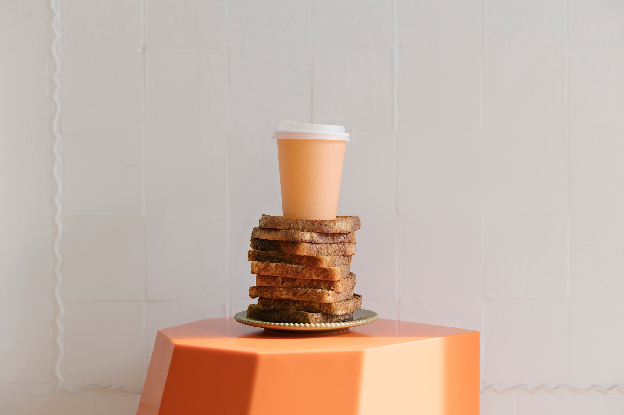 Landscape image of a orange hexagon shaped stool with a soft pink plate sitting in the middle. 12 pieces of wholemeal bread stacked on top, with an orange disposable cup placed on top. 