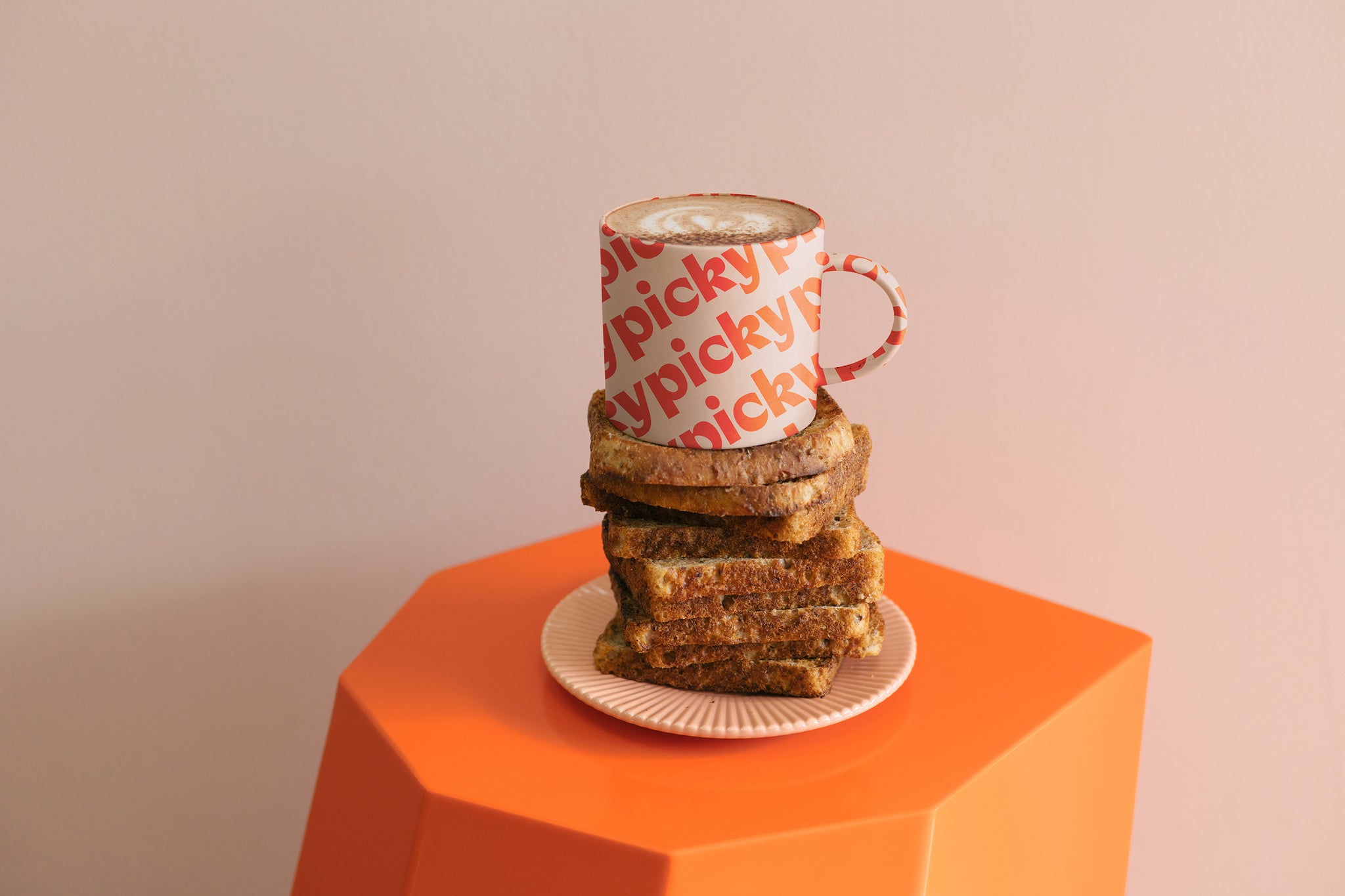 Landscape image of a orange hexagon shaped stool with a soft pink plate sitting in the middle. 12 pieces of wholemeal bread stacked on top, with a pink mug placed on top. The words Picky repeatedly displayed on an angle moving up the mug in a red colour. Three full words saying 'picky' can be read legibly. 