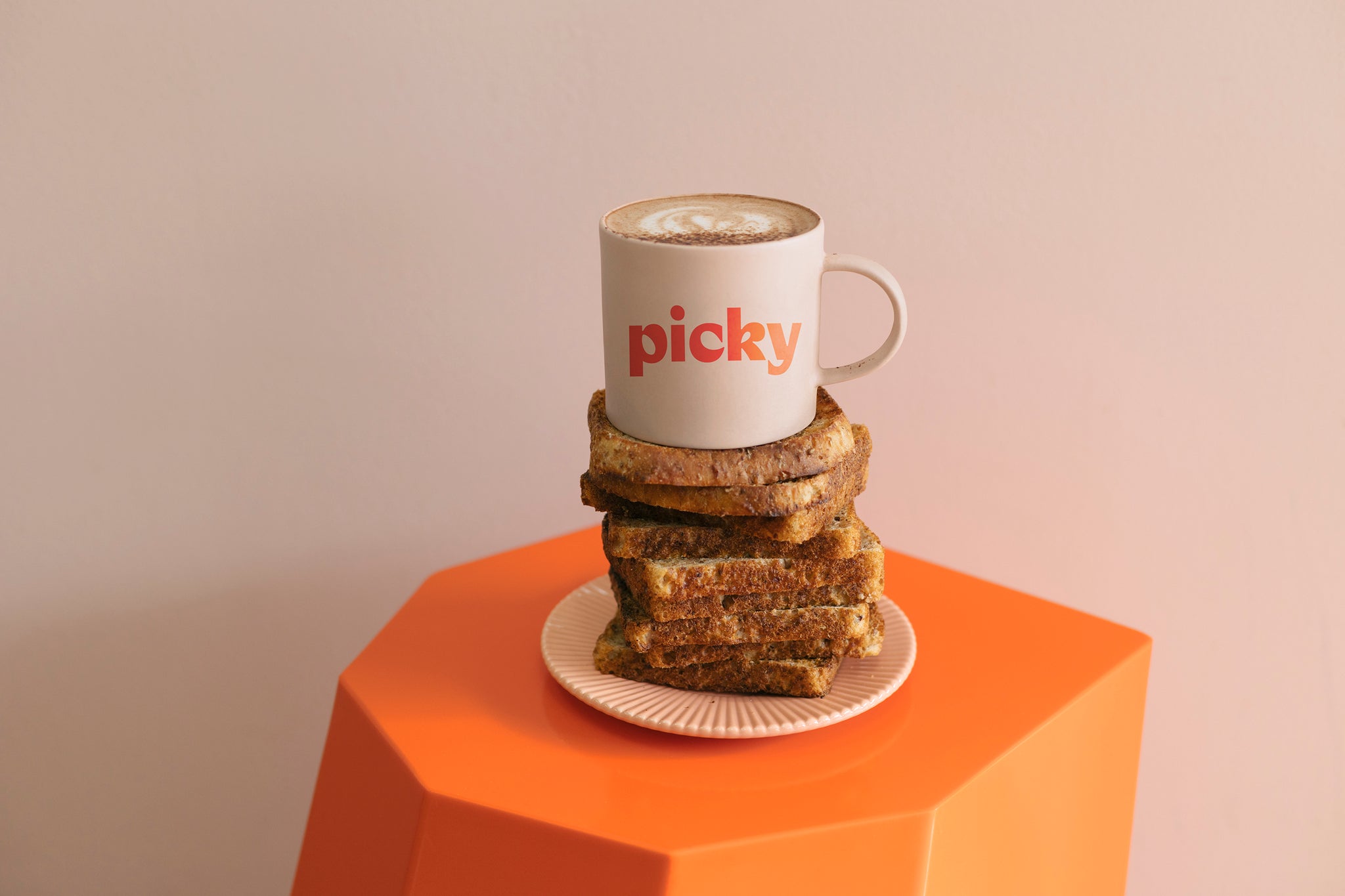 Landscape image of a orange hexagon shaped stool with a soft pink plate sitting in the middle. 12 pieces of wholemeal bread stacked on top, with a pink mug placed on top. The words Picky displayed straight acros the mug in a red colour. 