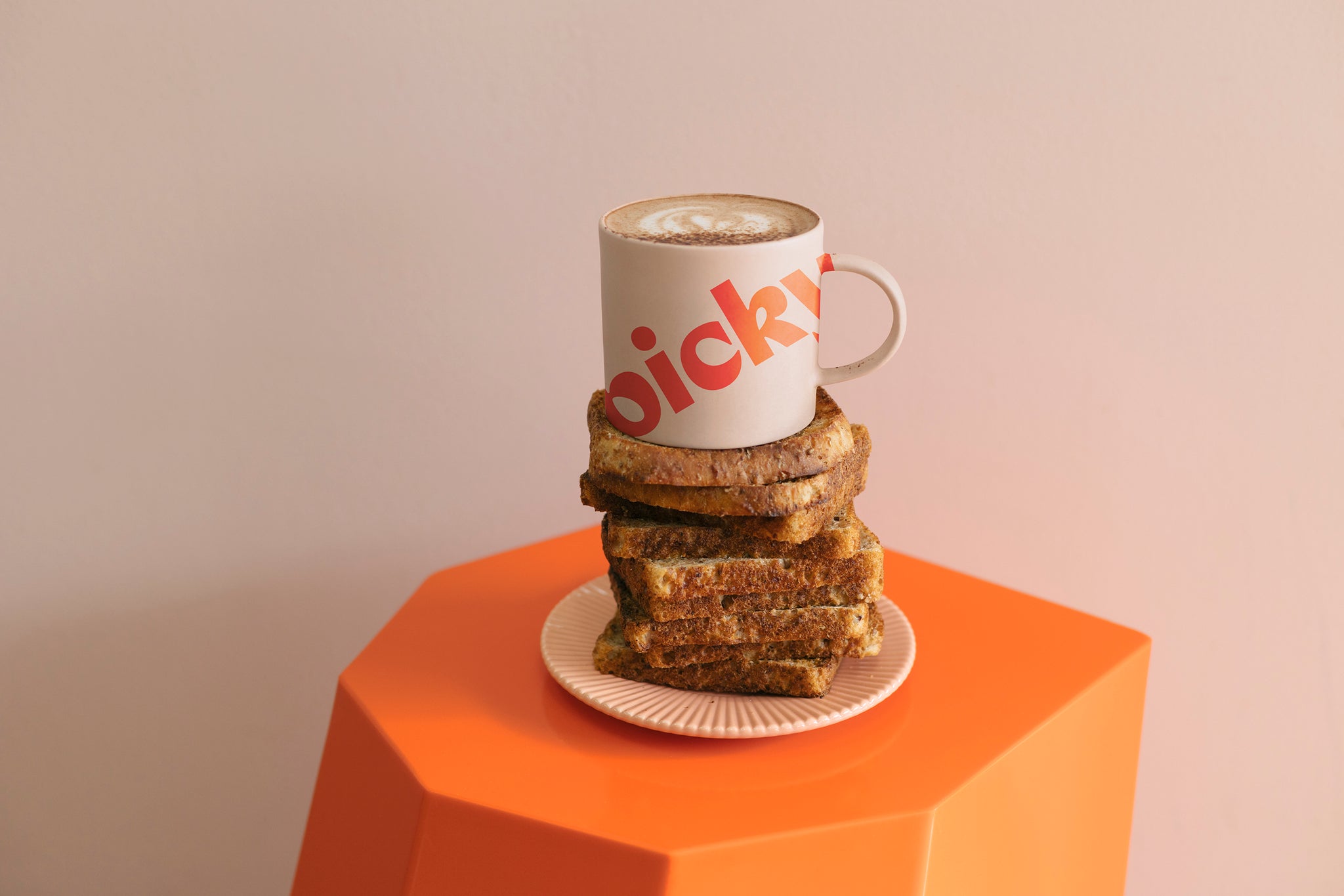 Landscape image of a orange hexagon shaped stool with a soft pink plate sitting in the middle. 12 pieces of wholemeal bread stacked on top, with a pink mug placed on top. The words Picky displayed largely on an angle moving up the mug in a red colour. 