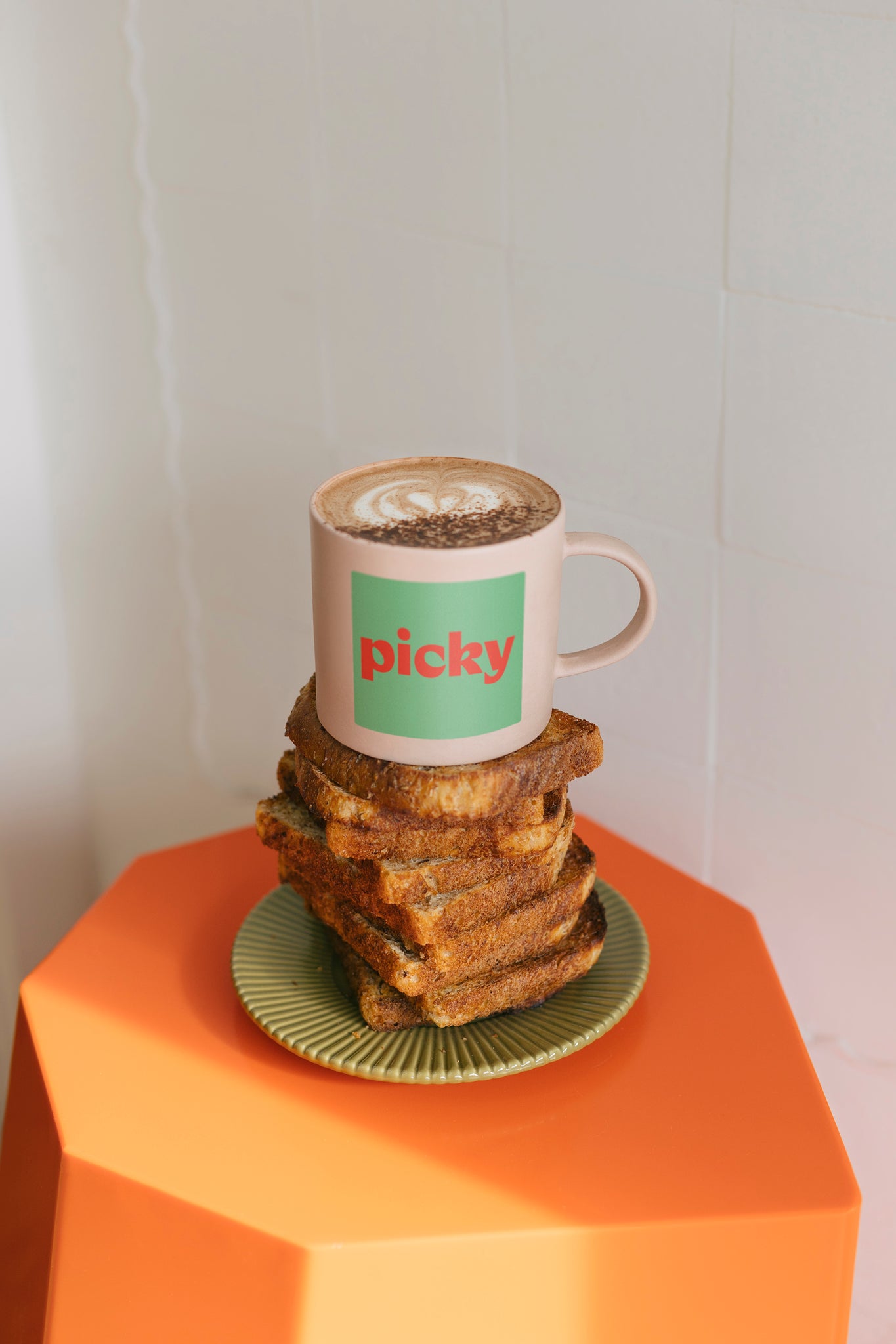 Portrait image of a orange hexagon shaped stool with a soft pink plate sitting in the middle. 12 pieces of wholemeal bread stacked on top, with a pink mug placed on top. The words Picky displayed inside a green box in the centre of the mug. 