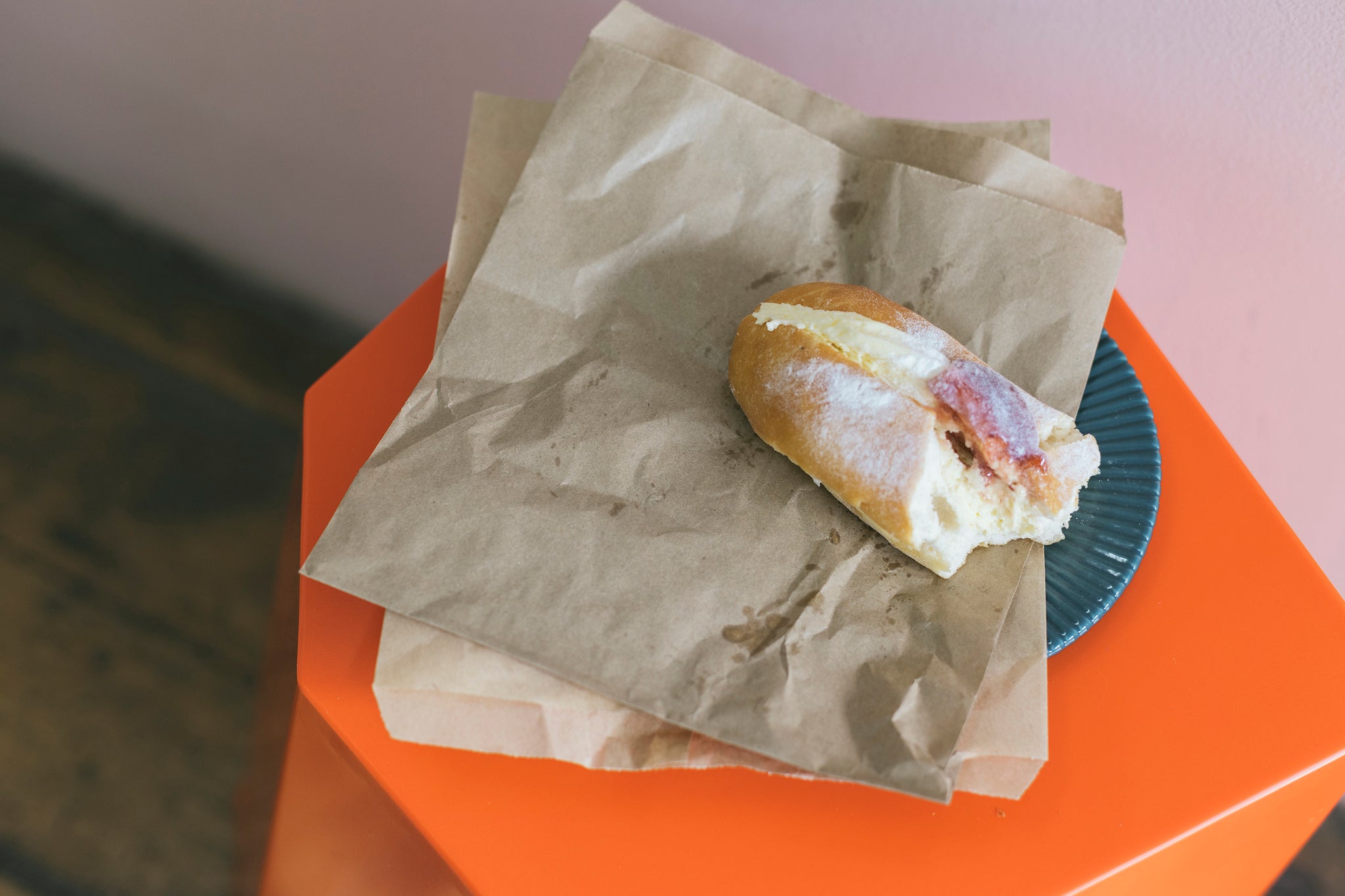 Brown paper bag placed on top of orange hexagon shaped stool. A Half eaten fresh cream donut with jam is place on top of bag. 