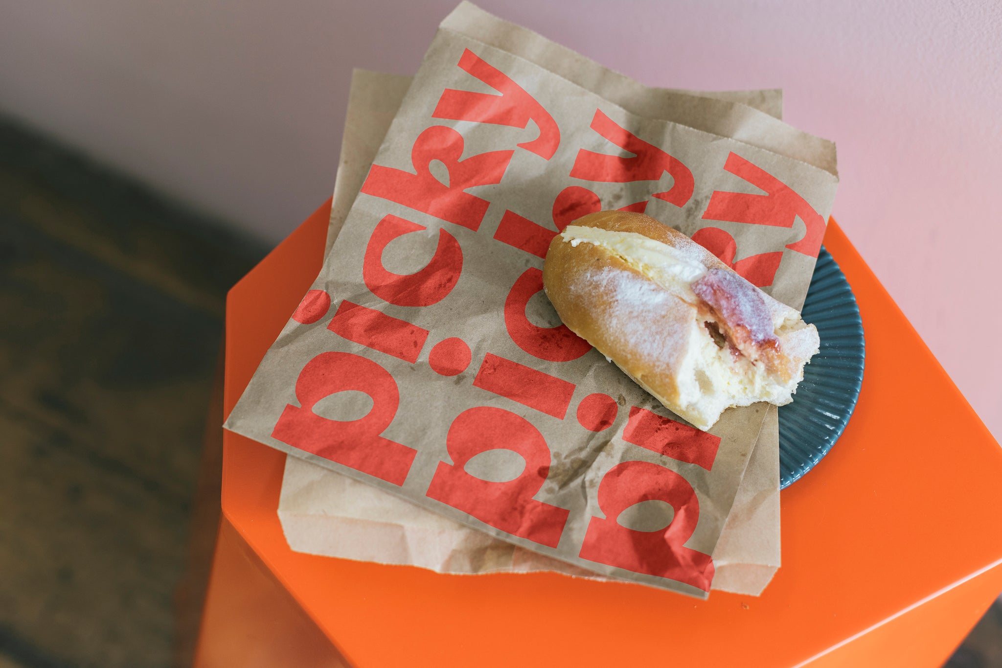 Brown paper bag placed on top of orange hexagon shaped stool. A Half eaten fresh cream donut with jam is place on top of bag. The words Picky is repeated three times in red on top of the bag. 
