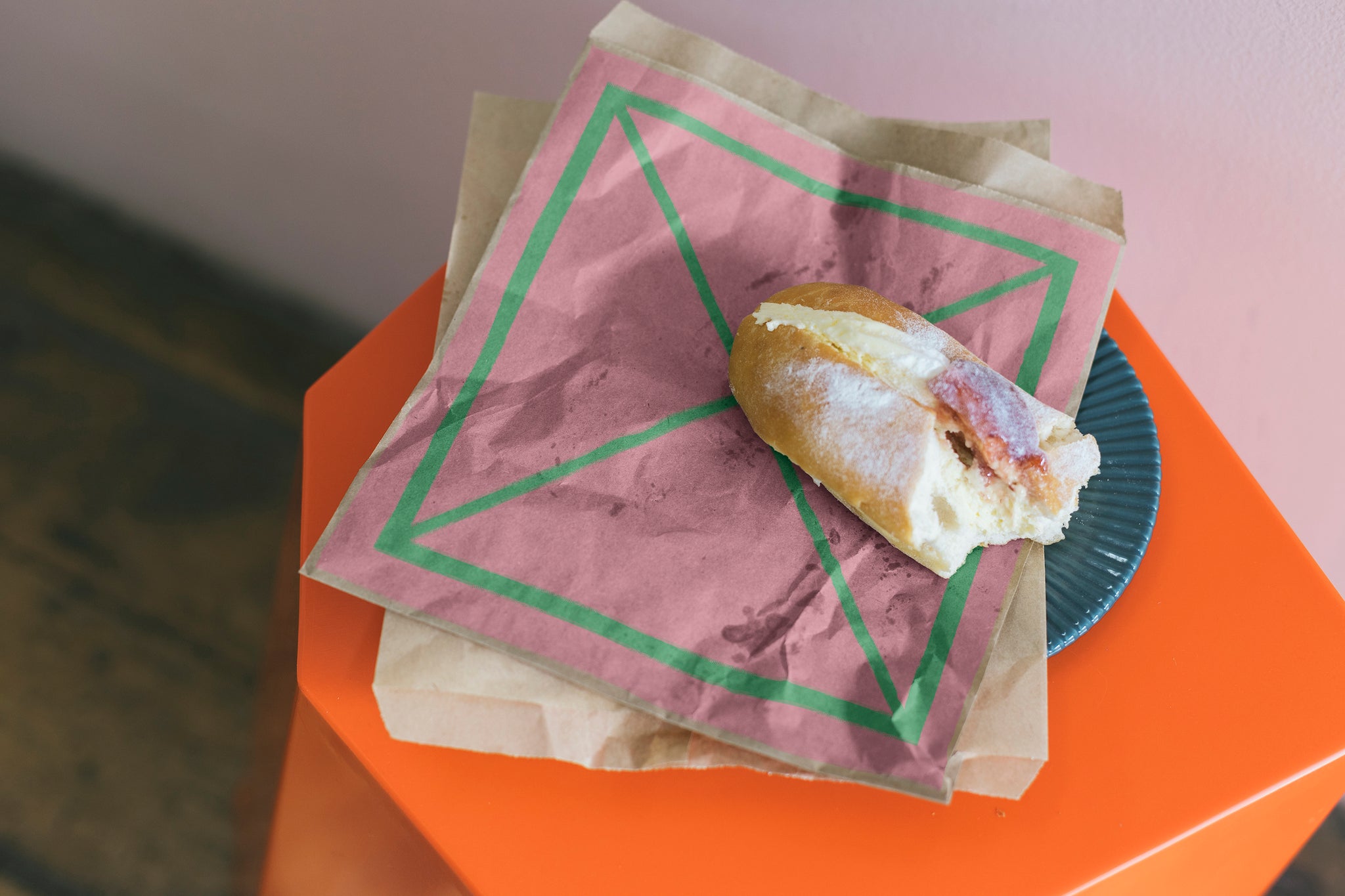 Brown paper bag placed on top of orange hexagon shaped stool. A Half eaten fresh cream donut with jam is place on top of bag. A big pink square is embedded on the bag with a bright green X in the middle of the square. 