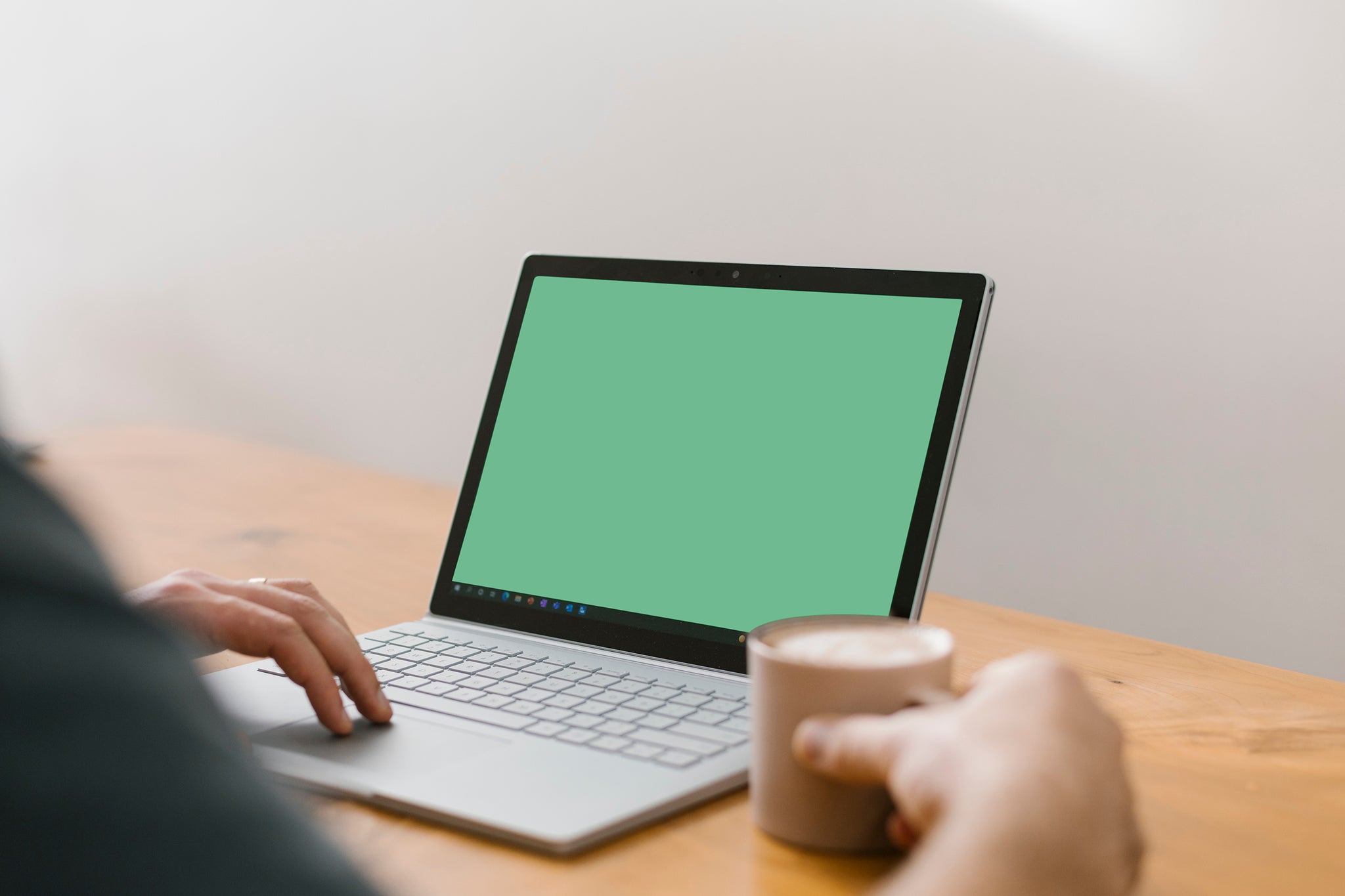 Landscape of an over-the-shoulder close-up of a person using a MacBook computer drinking a hot beverage. The monitor displays a plain green srcreen.