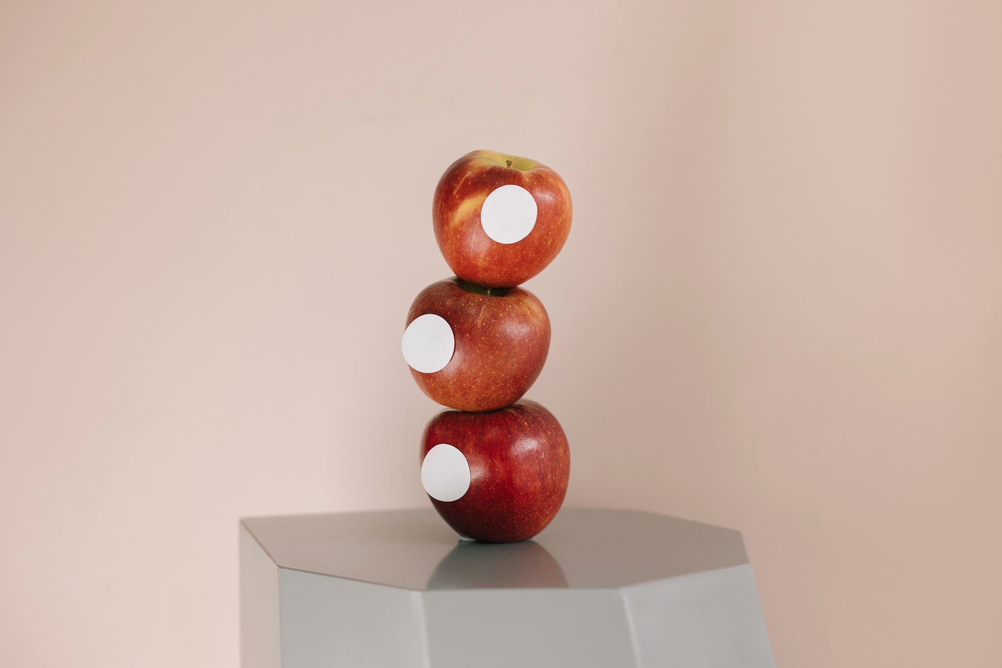 Landscape image of a grey hexagon shaped stool with three red apples stacked on top. Each apple with a white sticker placed on different parts. Backgoround is a soft pink. 