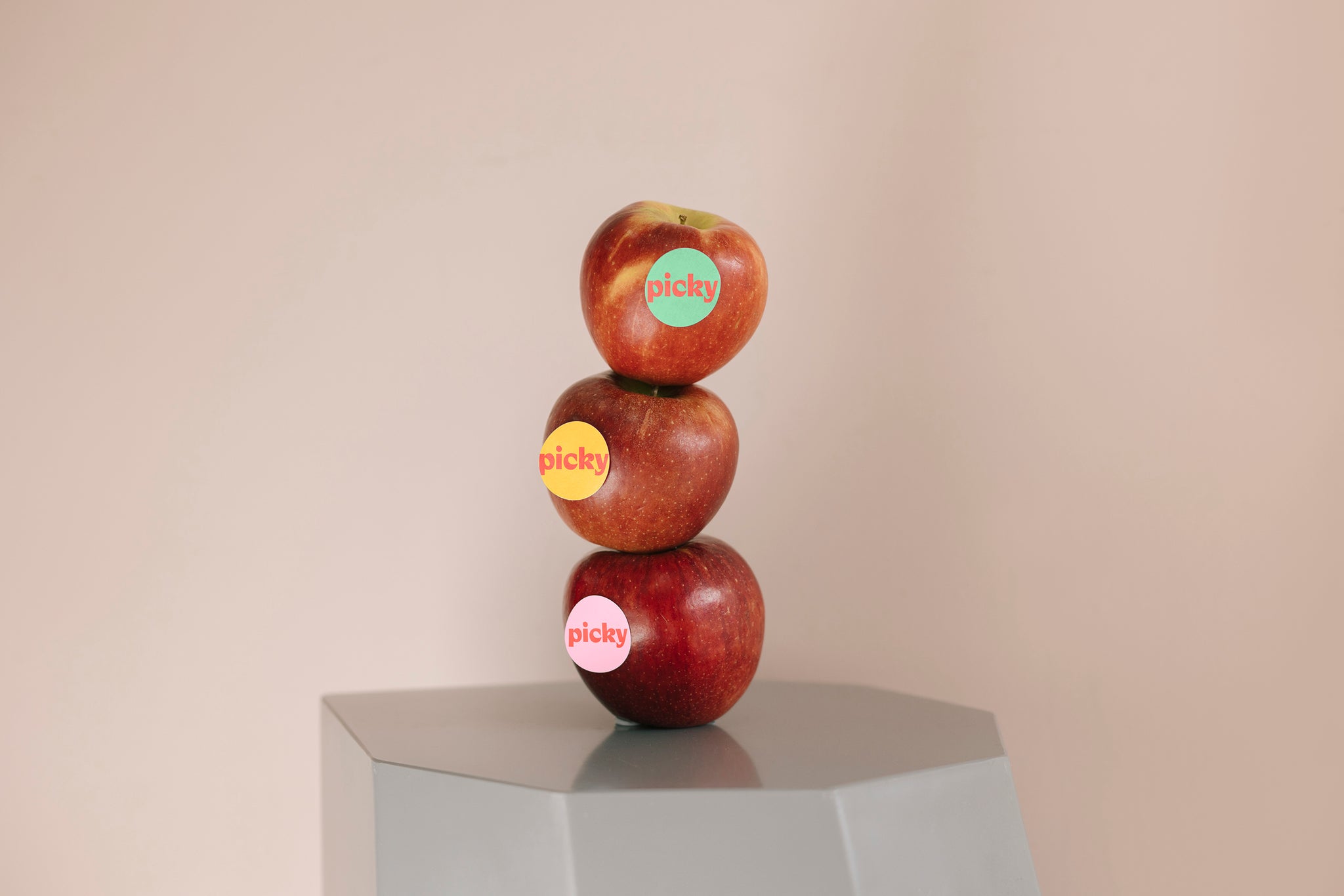 Landscape image of a grey hexagon shaped stool with three red apples stacked on top. Each apple with a sticker placed on different parts. The word Picky is written on each colouful sticker. Backgoround is a soft pink. 