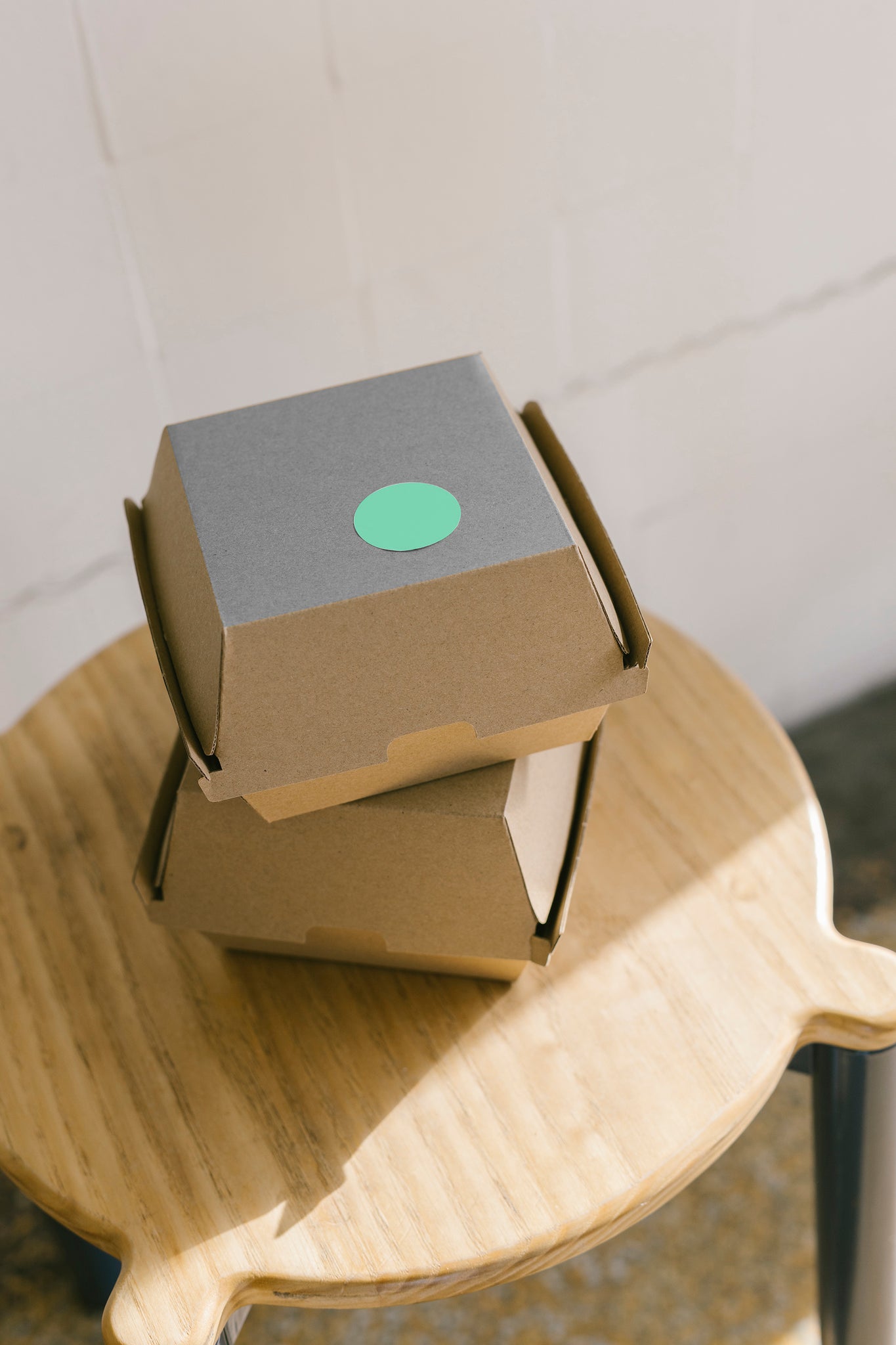 Portrait shot of two cardboard burger boxes stacked, sitting on top of a wooden stool. Light coming in from the window to the right of frame. A green sticker is stuck to the top box on the top side. 