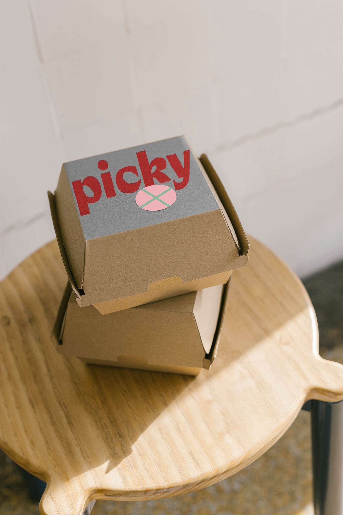 Portrait shot of two cardboard burger boxes stacked, sitting on top of a wooden stool. Light coming in from the window to the right of frame. The word Picky with a pink sticker is on the top of the first box. 