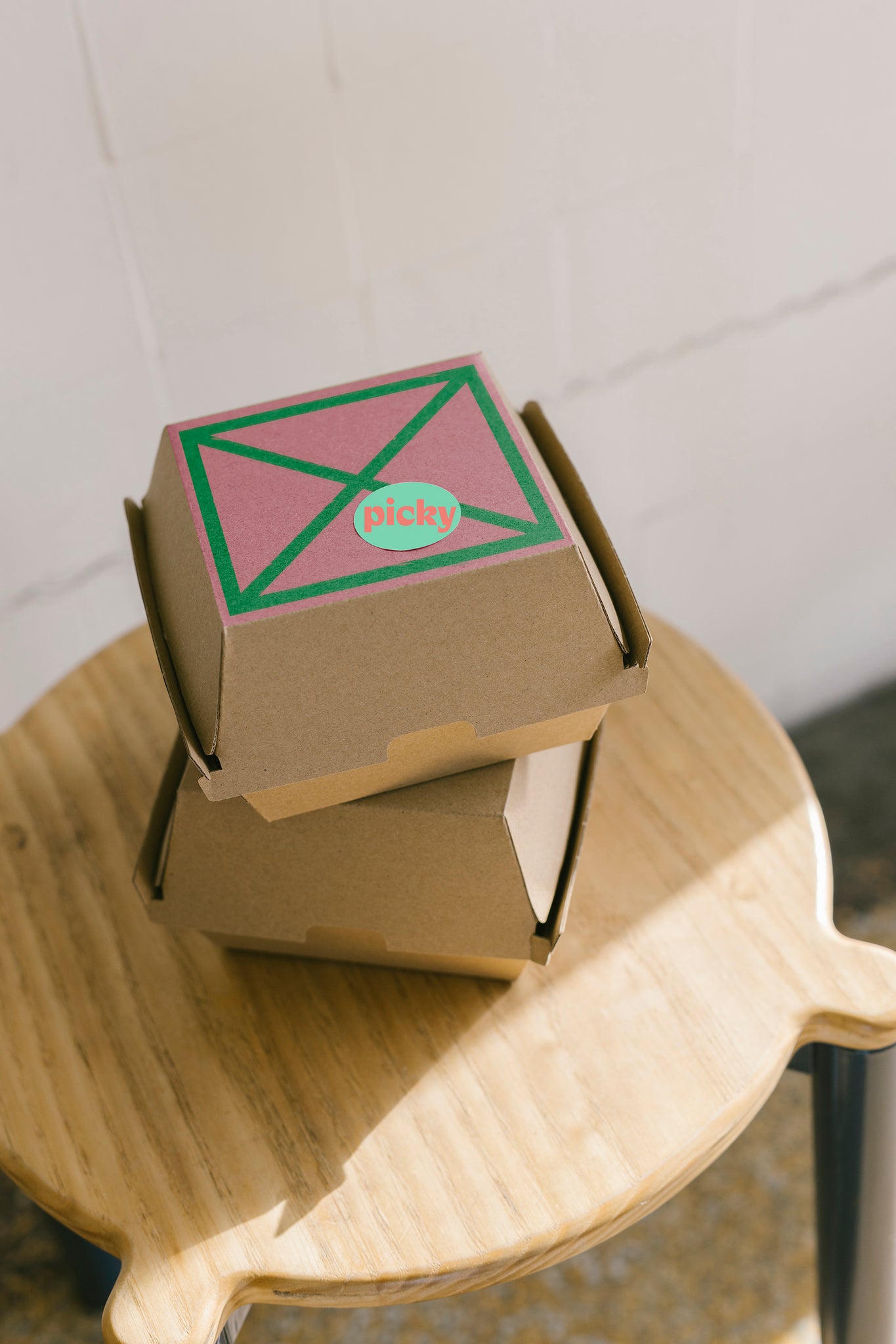Portrait shot of two cardboard burger boxes stacked, sitting on top of a wooden stool. Light coming in from the window to the right of frame. A pink square is framed on the top of the top burger box, with a green X in the middle of it. A green sticker is on top of the X with words Picky written in red. 