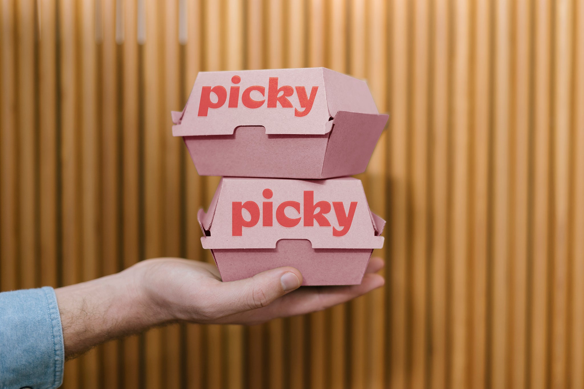 A white male hand with a visible blue denim sleeve is holding two pink cardboard burger boxes into the middle of the frame. Background is wooden poles. The word Picky is written in red on the front face of each box. 