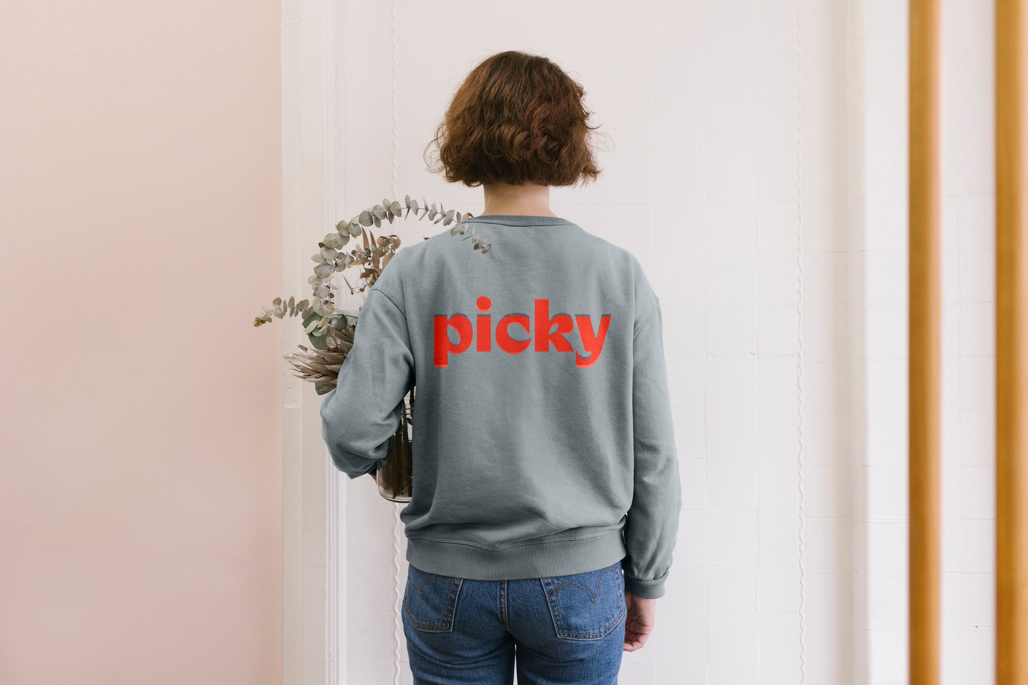 A lady is standing in front of a wall holding a eucalyptus plant. She is wearing a grey sweatshirt with the word Picky written on the back, written in red and placed in the top middle. Her hair is brown and sits at the height of her ears. The background is a soft pink.