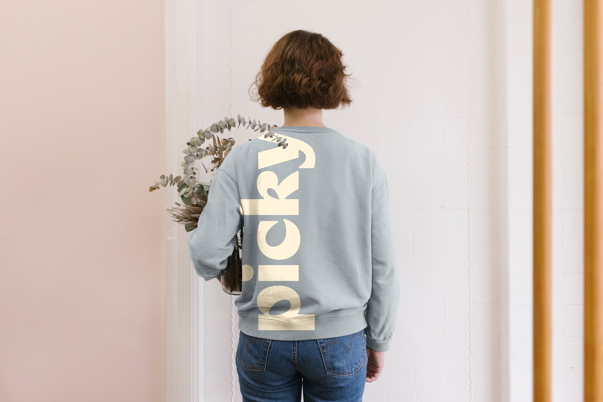 A lady is standing in front of a wall holding a eucalyptus plant. She is wearing a grey sweatshirt with the word Picky written on the back, written from the left hand side towards her shoulder. Her hair is brown and sits at the height of her ears. The background is a soft pink.