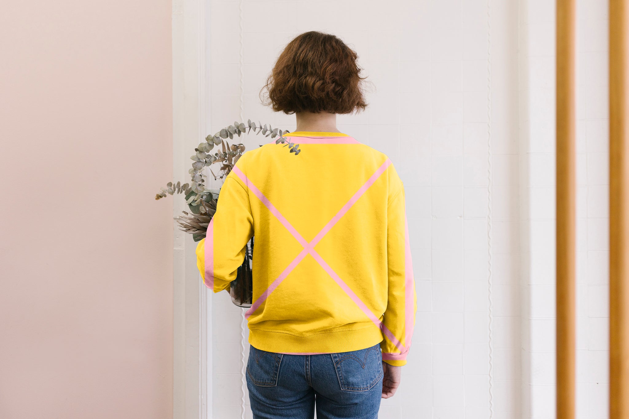 A lady is standing in front of a wall holding a eucalyptus plant. She is wearing a yellow sweatshirt with a large pink X grid line on her back. Her hair is brown and sits at the height of her ears. The background is a soft pink.