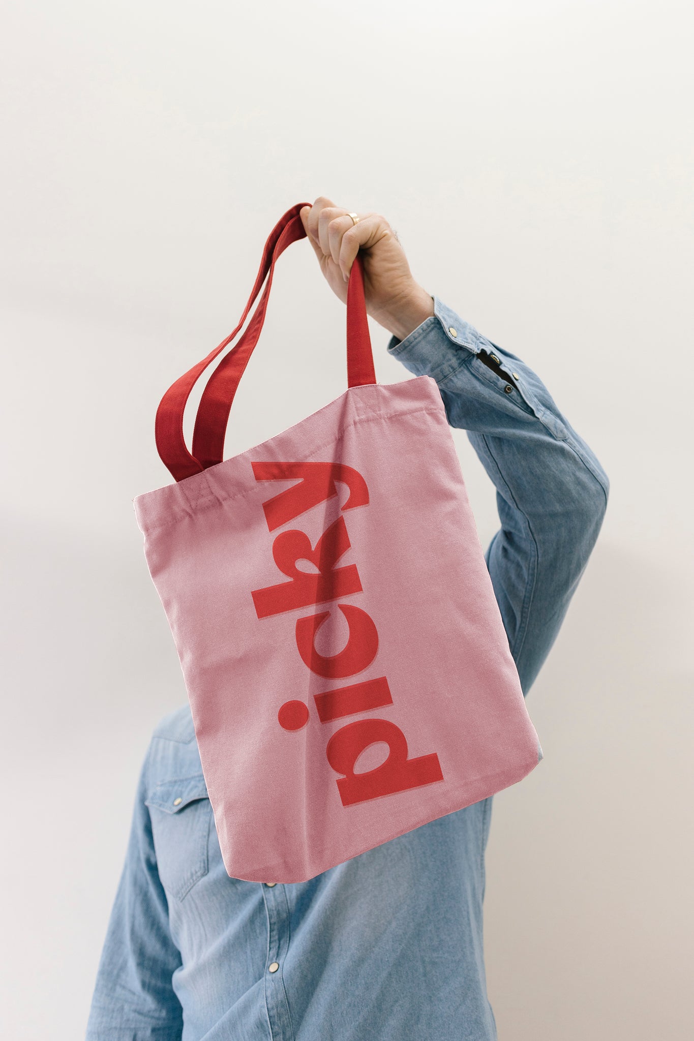 Person holding a pink tote bag by its handles positioned over their head, covering themselves. Bag has the word Picky noted in a red in the horizontal in the middle of the bag. Person is standing against a plain grey wall wearing a long sleeve denim shirt. 