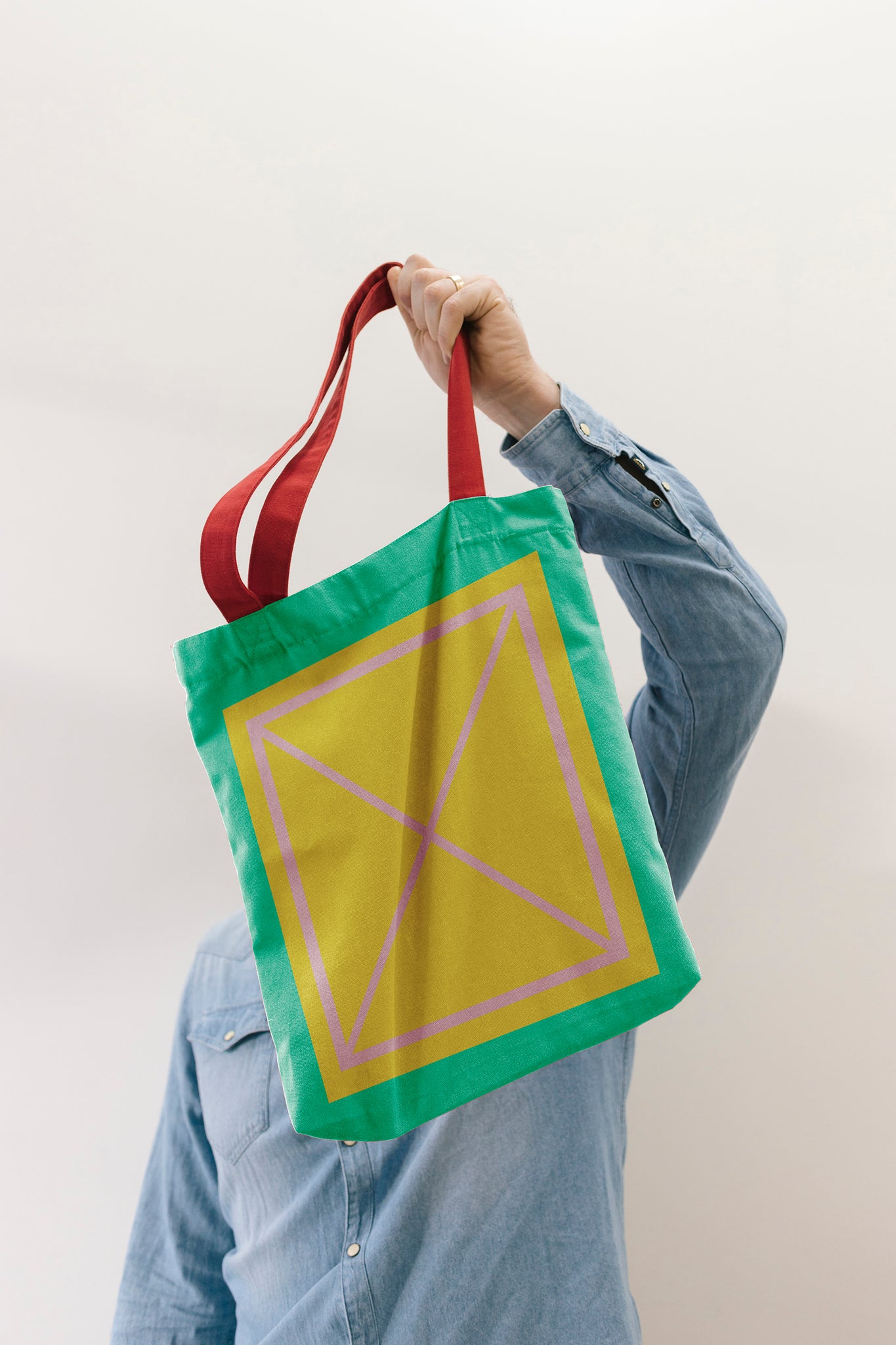 Person holding a green tote bag by its red handles positioned over their head, covering themselves. Bag has a large yellow box in the middle of the bag, with the pink X grid line in the middle. Person is standing against a plain grey wall wearing a long sleeve denim shirt. 