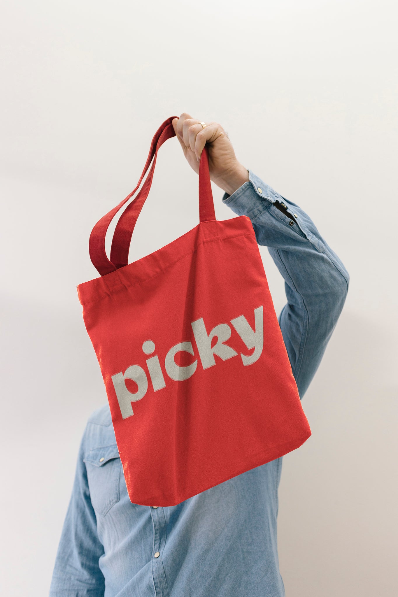 Person holding a red tote bag over their head. Bag has the word Picky noted in a soft white colour in the middle of the bag. Person is standing against a plain grey wall wearing a long sleeve denim shirt. 