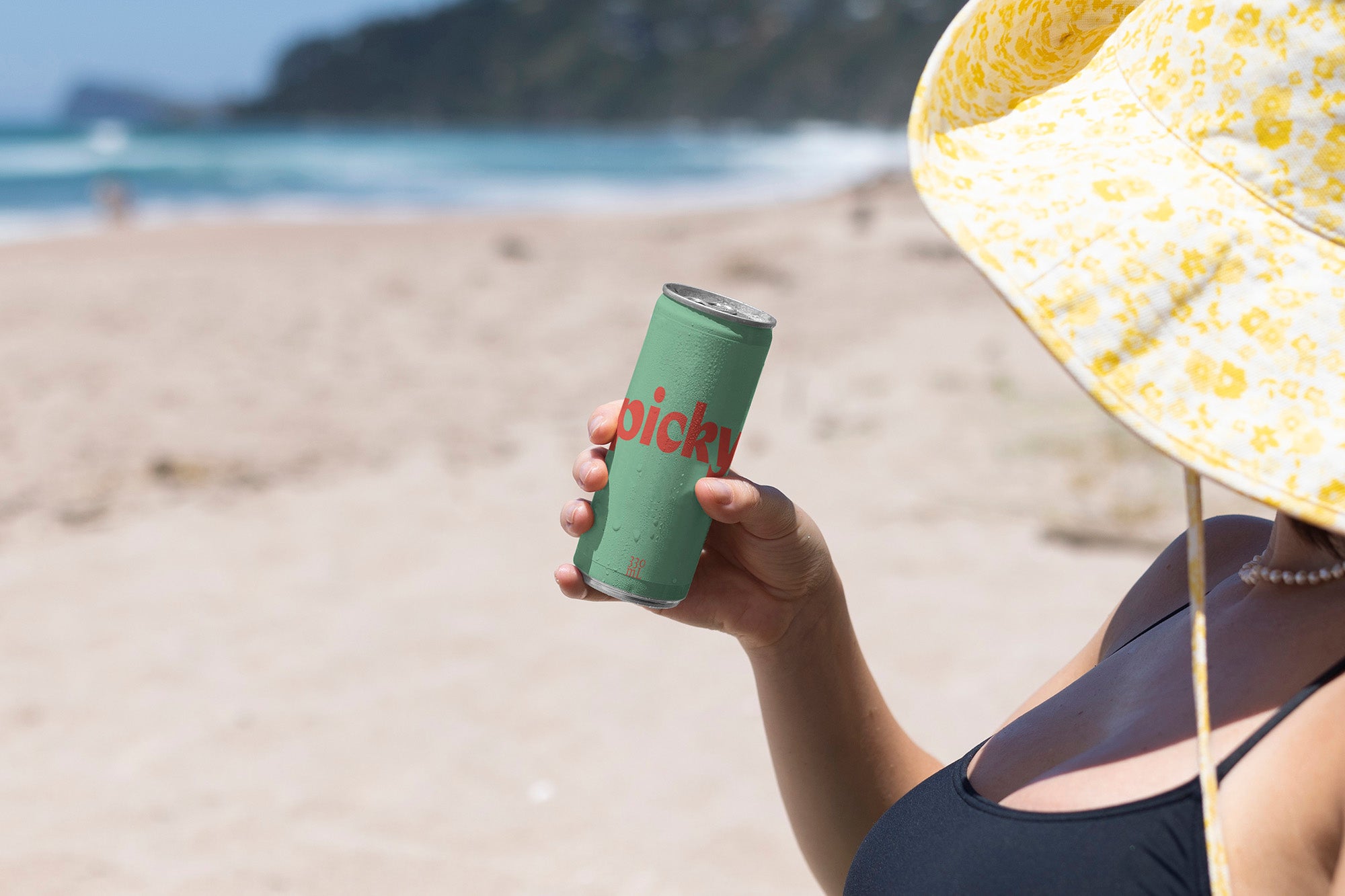 Lady with large brim hat holds bright coloured slim drinks can on beach.