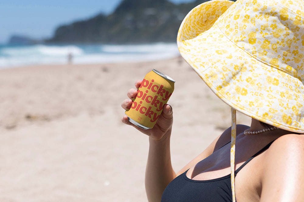 Lady with large brim hat holds bright coloured drinks can on beach.
